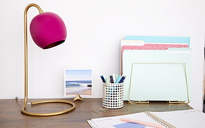 A desk with a calendar, file folders, jar of pens and a lamp with a gold base and magenta shade.