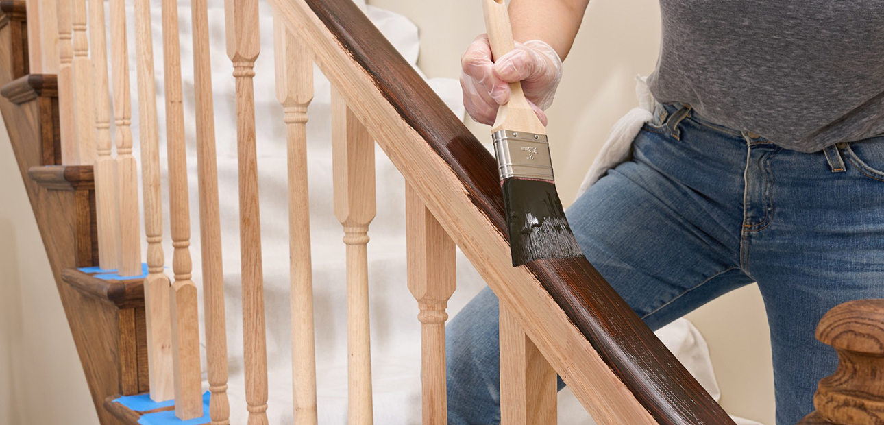 Using a brush to apply gel stain to a wooden staircase railing.