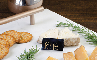 Rose gold handles on a white serving tray that has crackers, pieces of brie and gouda cheese and garnishes on top.
