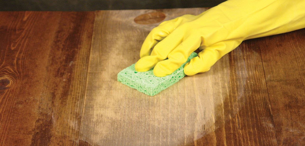 A gloved hand applying wood bleach with a sponge. 