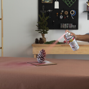 A woman applying red spray paint to a pine cone. 