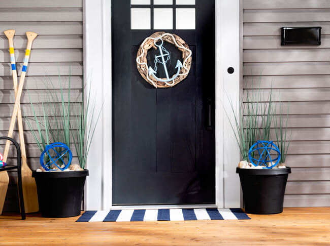 A nautical-themed front porch with an anchor wreath on the front door, urn planters filled with blue orbs and tall green grass and two wooden oars. 