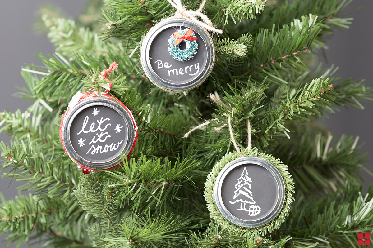 Three chalkboard jar lid ornaments with let it snow, be merry and a Christmas tree drawn on them in chalk. 