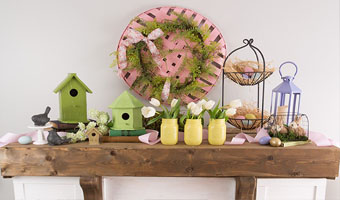 A wooden mantel covered with pastel colored spring décor, including small wooden birdhouses, yellow vases filled with tulips, a two-tiered basket filled with Easter eggs and a spring wreath with a pink bow above the mantel.