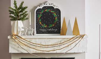 Silver and gold beaded garland in front of a mantel that’s decorated with evergreens in a vase, small white reindeer, a large sign that reads Happy Holidays and silver and gold wooden decorative trees.